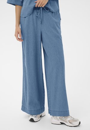 Personne portant un pantalon en denim bleu clair ample avec une taille à cordon de serrage et des baskets blanches New Balance sur un fond blanc.