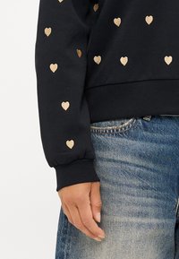 Un sweat-shirt noir avec des motifs de cœurs dorés, des poignets et un ourlet côtelés. Assorti avec un jean bleu, mettant en avant un style décontracté.