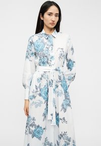 Φλοράλ φόρεμα με λευκό φόντο, διακοσμημένο με μπλε και γκρι τριαντάφυλλα. Μακριά μανίκια, γιακάς με κουμπιά και ζώνη δεμένη στη μέση.