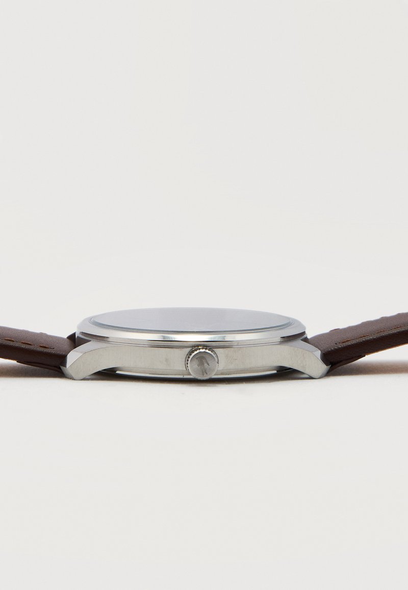 Silberne Armbanduhr mit rundem Zifferblatt, braunem Lederarmband und seitlicher Krone, seitlich auf weißem Hintergrund dargestellt.