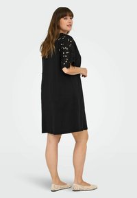 Čierne šaty s krátkymi, vlnitými čipkovými rukávmi, vo voľnom strihu a po kolená. Modelka má na sebe svetlé, vzorované balerínky.