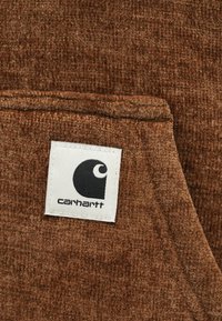 Tecido texturado castanho com um rótulo quadrado branco costurado, apresentando um logótipo Carhartt preto e o nome da marca.