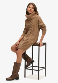 Beiges Strickkleid mit hohem, gefalteten Kragen und strukturierten Zopfmustern, kombiniert mit braunen kniehohen Stiefeln mit Schnallen.