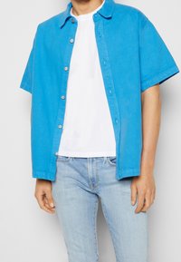 Camicetta a maniche corte con bottoni in cotone blu brillante, dal fit rilassato. Presenta un colletto e bottoni argentati, indossata sopra una t-shirt bianca.