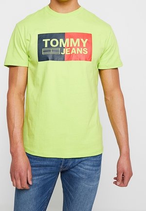Mężczyzna w jasnorozowej koszulce z krótkim rękawem z logo Tommy Jeans w czarnym, czerwonym i żółtym na piersi, w połączeniu z niebieskimi dżinsami.