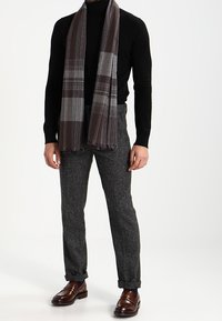 Col roulé noir, écharpe en plaid gris avec des bords frangés, pantalon texturé gris et chaussures en cuir marron. Mettre l'accent sur les matériaux et les couleurs.
