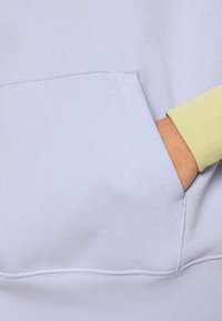 Ljus lavendelfärgad sweatshirt med en känguruficka och kontrasterande ljusgul muddstycke. Mjuk textur med synliga sömdetaljer.