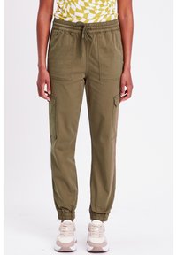 Pantaloni jogger verde oliva con vita elastica, due tasche frontali e polsini a coste. Indossati con scarpe da ginnastica crema e rosa.