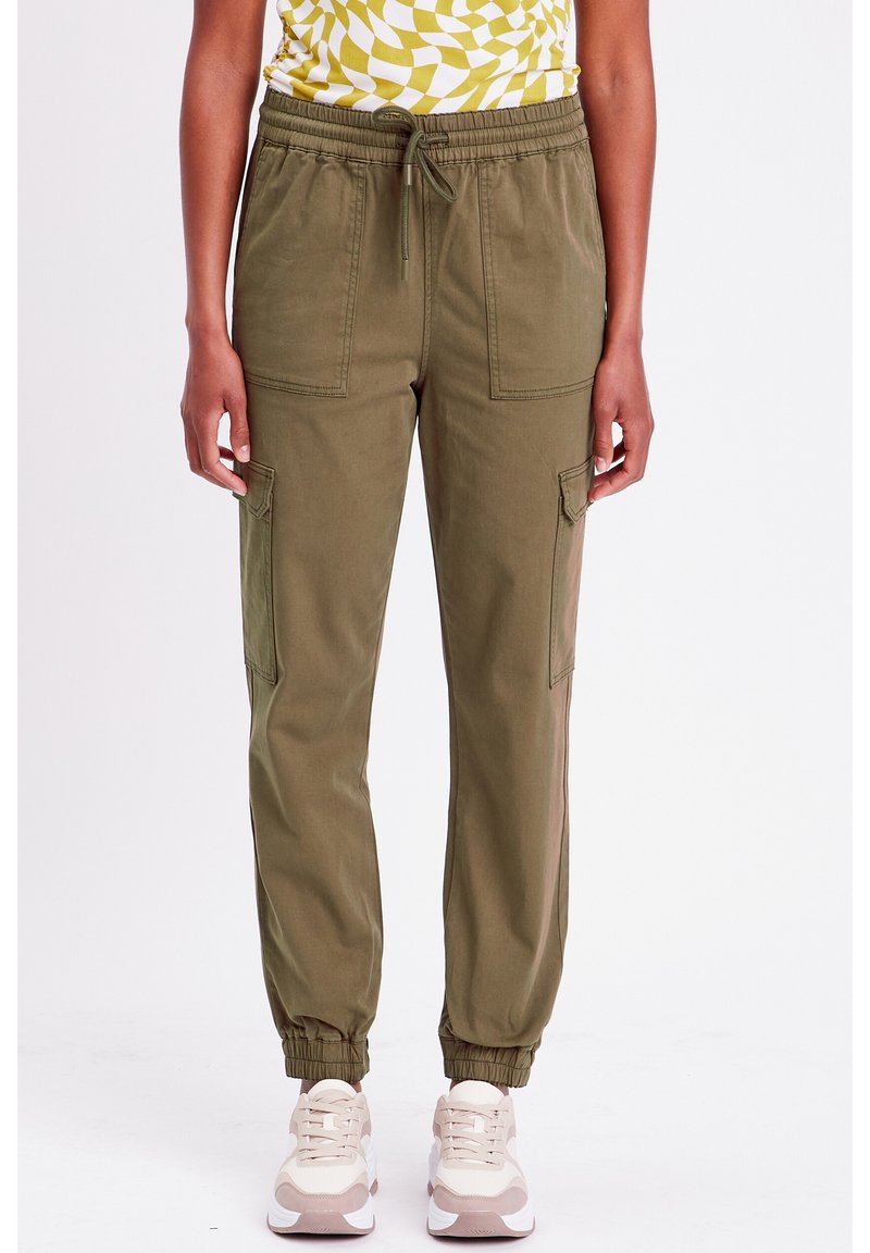 Pantalons de jogger vert olive avec une taille élastique, deux poches avant et des poignets côtelés. Portés avec des chaussures de sport crème et rose.