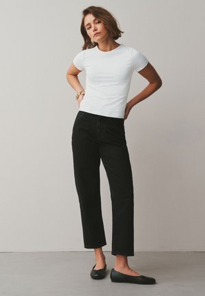 Frau steht mit den Händen hinter dem Rücken, trägt ein weißes Kurzarmshirt, schwarze weite Hosen, schwarze Ballerinas und ein goldenes Armband.