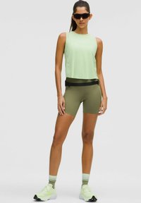 Giovane donna che indossa un top senza maniche verde chiaro, pantaloni biker verde oliva, sneaker verde neon e occhiali da sole neri, in piedi contro uno sfondo semplice.