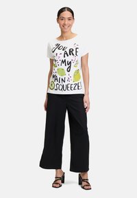 Biała bawełniana koszulka z wyrazistym czarnym napisem i kolorowymi grafikami owoców. Połączona z czarnymi szerokimi spodniami i czarnymi sandałami na obcasie.