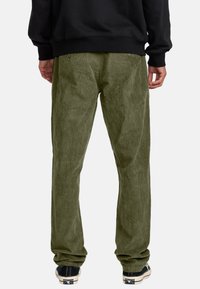 Olive green corduroy pants with a textured surface, straight cut, and black sneaker accents. Details include two back pockets with buttons.