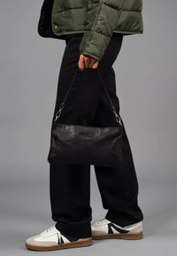Bolso de piel negra con una textura suave, forma rectangular delgada y correa corta, sostenido por una persona que lleva una chaqueta acolchada verde y pantalones negros.
