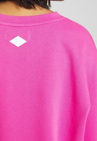 Camiseta de algodón rosa de gran tamaño con cuello redondo, que presenta un acento en forma de diamante blanco en la parte posterior. Textura suave y corte relajado.