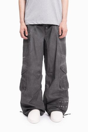 Osoba nosząca luźne ciemnoszare spodnie cargo z wieloma kieszeniami i metalowymi oczkami, w połączeniu z jasnoszarym koszulą i białymi butami.