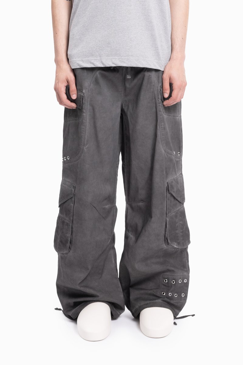 Osoba nosząca luźne ciemnoszare spodnie cargo z wieloma kieszeniami i metalowymi oczkami, w połączeniu z jasnoszarym koszulą i białymi butami.