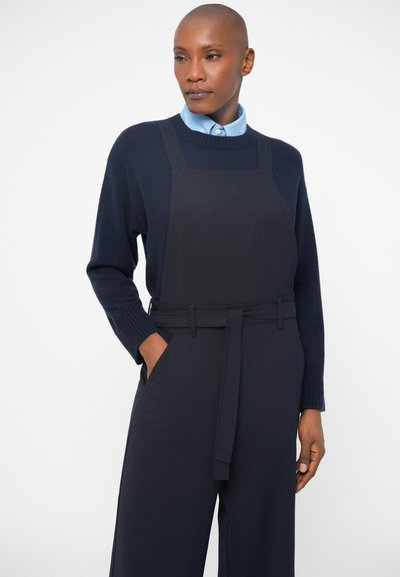 Džemper v mornarsko modri barvi iz pletene tkanine, v kombinaciji z enobarvno črno obleko z predpasnikom, ki ima pas z dodano širino, široke hlače in stranske žepke. Gladka tekstura tkanine.