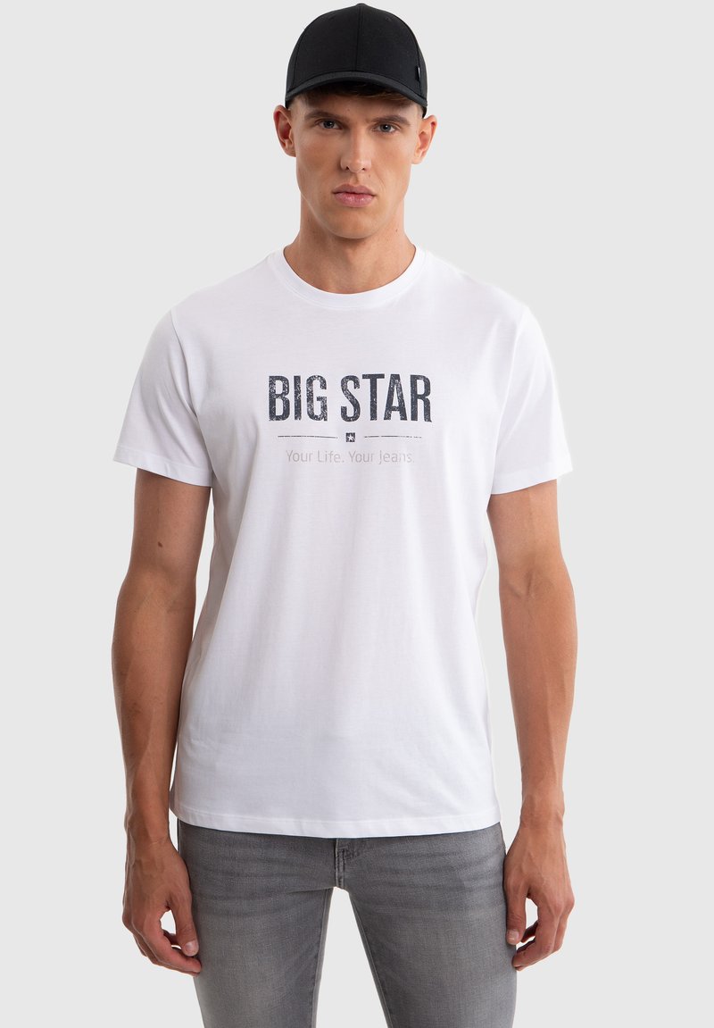 Uomo con cappellino nero, maglietta bianca con la scritta "BIG STAR Your Life. Your Jeans" e jeans grigi, in piedi davanti a uno sfondo chiaro e semplice.