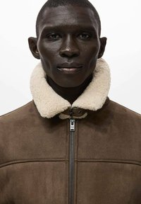 Giacca in suede marrone con colletto in eco-pelliccia color crema, chiusura con zip e dettagli cuciti. Texture liscia con un design moderno.