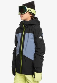 Giacca da sci nera e blu con una cerniera e dettagli verdi, dotata di cappuccio, tasche con zip e guanti coordinati; indossata con occhiali da sci.