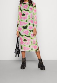 Robe à manches longues en tissu léger et extensible, présentant un motif abstrait rose, vert et noir avec des fentes sur les côtés. Associée à des bottes noires imposantes.