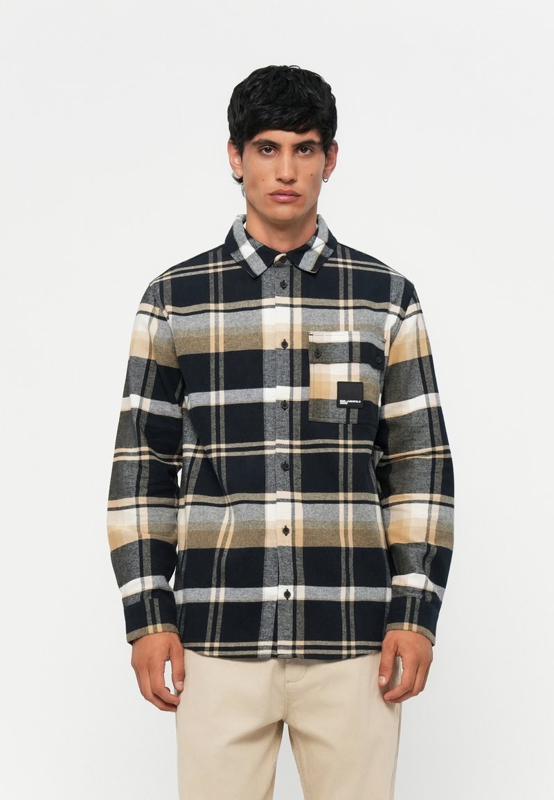 Camicia di flanella con motivo a quadri nei colori nero, giallo, grigio e bianco. Ha chiusura con bottoni, colletto e taschino sul petto a sinistra. Texture morbida.