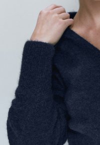 Person iført en tekstureret mørkeblå fuzzy trøje med V-hals, hånd hviler nær kravebenet mod en ensfarvet lys baggrund.