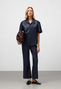 Kobieta w granatowej koszuli z krótkim rękawem i dopasowanych szerokich spodniach, trzymająca ciemnoczerwoną skórzaną kopertówkę, stojąca na szarym podłożu.