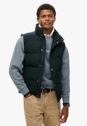 Gilet imbottito nero con colletto alto sopra una felpa azzurra. Pantaloni khaki marroni. Accentuato da bottoni metallici e una cerniera.