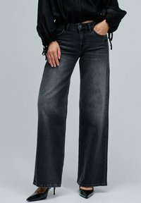 Mujer con jeans anchos de tiro alto negros, blusa negra de manga larga con puños atados y tacones altos puntiagudos negros, de pie frente a un fondo gris.