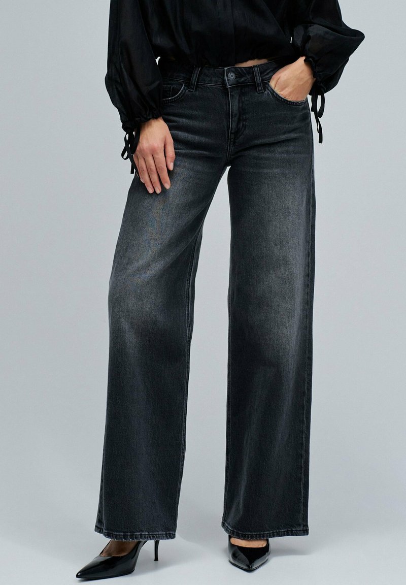 Mujer con jeans anchos de tiro alto negros, blusa negra de manga larga con puños atados y tacones altos puntiagudos negros, de pie frente a un fondo gris.