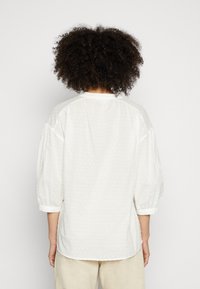 Blusa branca com bolinhas texturizadas, corte solto, mangas bufantes e decote redondo. O tecido parece leve e respirável.