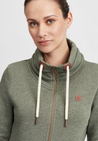 Sidelag jakke i olivengrønn, med høy krage, beige snorer og brune detaljer. Mykt materiale med en glatt tekstur.