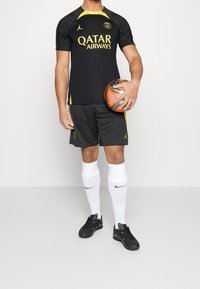 Camiseta deportiva negra con acentos amarillos, con el logo de "Qatar Airways". Pantalones cortos negros y calcetas blancas hasta la rodilla. Sosteniendo un balón de fútbol naranja.