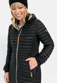 Zwarte pufferjas met gewatteerde textuur, ritssluiting en capuchon. Heeft twee zijzakken, geribbelde manchetten en een bruine leren accent.
