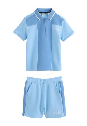 Junger Junge, der lächelt und steht, trägt ein hellblaues kurzärmeliges Poloshirt, passende Shorts, weiße Socken und weiße Turnschuhe.