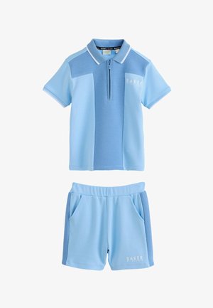 Ung dreng, der smiler og står, iført en lyseblå kortærmet polo, matchende shorts, hvide sokker og hvide sneakers.