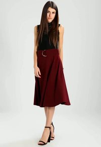 Saia midi bordô com cintura cinguada, apresentando um detalhe em anel de metal, combinada com uma blusa preta sem mangas e sandálias de salto pretas.