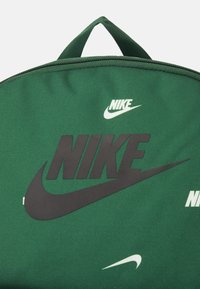 Grön Nike-ryggsäck med texturerad tyg, stort svart logotyp och mindre vita logotypdetaljer, med ett handtag på toppen och dragkedja.