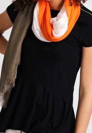 Femme portant un haut noir à manches courtes avec une écharpe tricolore orange, blanche et vert olive portée lâchement sur une épaule.