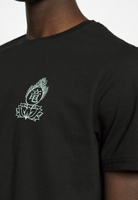 Černé bavlněné tričko s mátově zelenou grafikou lotosového květu a stylizovanými akcenty ve středové části na hrudi.