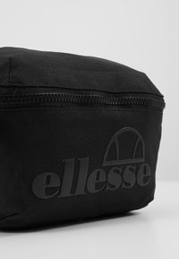 Trousse de toilette en tissu noir avec une texture lisse, dotée d'une fermeture éclair sur le dessus et d'un logo en relief sur le côté. Design rectangulaire simple.