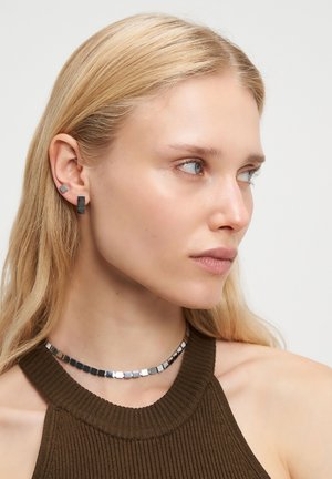 Geometriske øreringe har en blanding af rektangulære og kvadratiske former i sorte og metalliske nuancer. En kraftig choker med firkantet mønster omkranser halsen.
