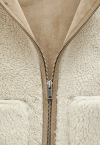 Gros plan d'une veste beige avec une fermeture éclair métallique partiellement ouverte montrant une doublure en sherpa polaire blanche douce et un tissu extérieur ressemblant à du daim.