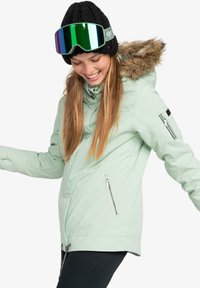 Mint green ski jacket with fur-lined hood, front zippers, and side pockets. Paired with a black knitted hat and reflective ski goggles.