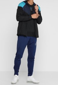 Schwarze Zip-Jacke mit blauen Akzenten, kombiniert mit marineblauen, schmal zulaufenden Hosen und weißen Sportschuhen. Besitzt einen glatten, leichten Stoff.