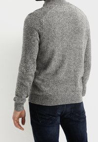 Grauer, gestrickter Pullover mit hohem Kragen, strukturiertem Muster und langen Ärmeln, kombiniert mit dunklen Jeans und sichtbaren Nähdetails.