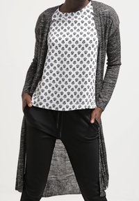 Larga chaqueta gris sobre una blusa blanca con un diseño estampado que presenta flores negras, combinada con pantalones holgados negros. Tela suave y texturizada.