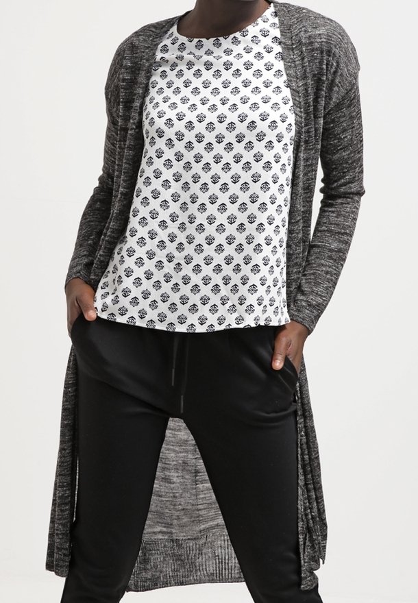 Larga chaqueta gris sobre una blusa blanca con un diseño estampado que presenta flores negras, combinada con pantalones holgados negros. Tela suave y texturizada.
